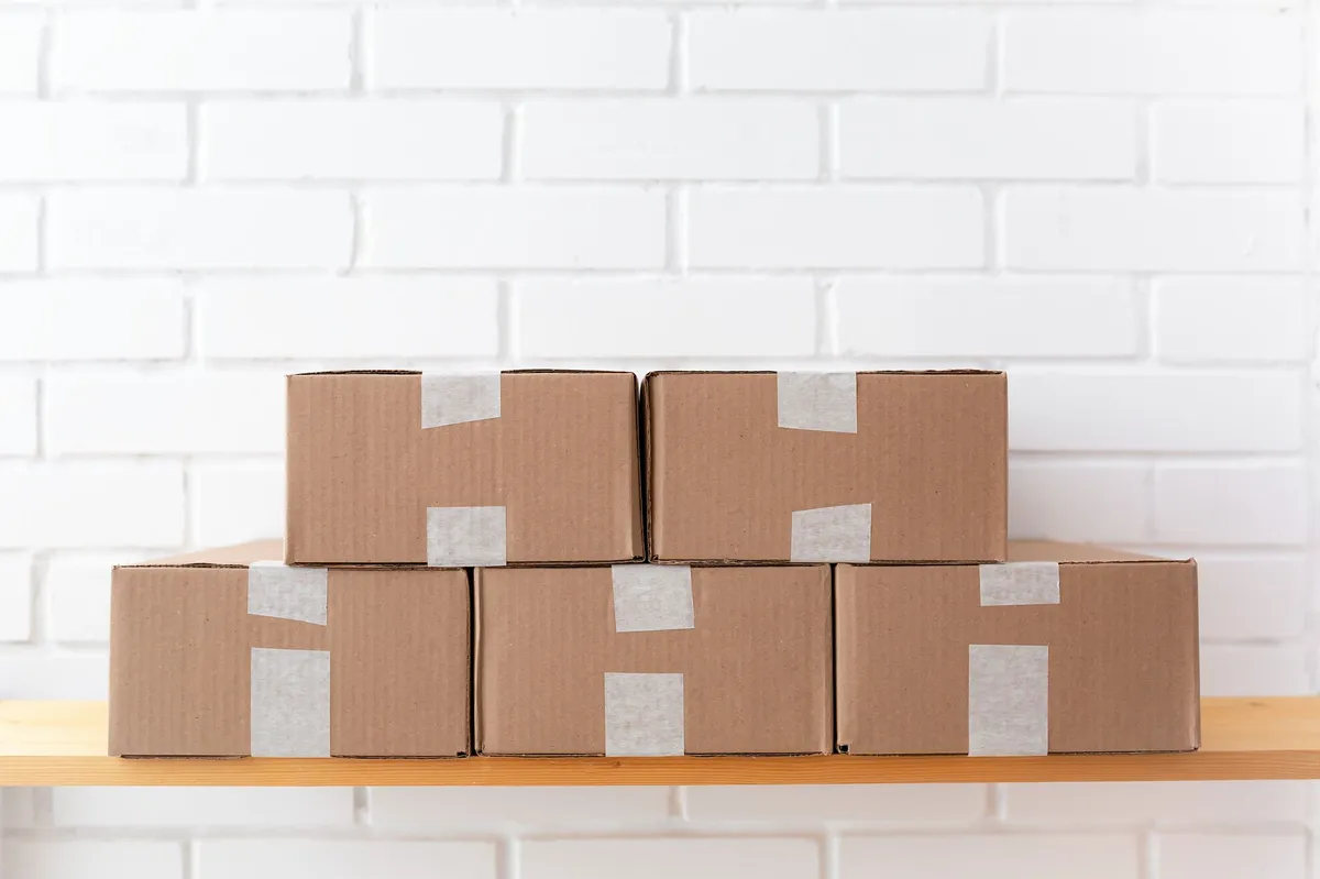 Cinco cajas de cartón selladas con cinta, apiladas sobre un estante de madera contra una pared blanca.